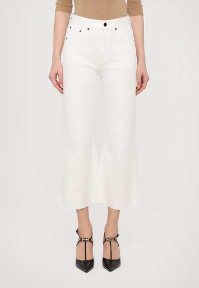 Jean large blanc en denim, avec un ourlet effiloché, un design à cinq poches et des boutons noirs. Porté avec des talons noirs à bride cheville.