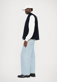 Gilet en polaire marine sur un sweat à capuche blanc, associé à un jean large bleu clair et des bottes noires. Une casquette blanche finalise la tenue.