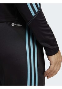 Černá sportovní bunda s modrými pruhy, vyrobená z prodyšného materiálu AEROREADY a s těsnými manžetami. Detailní záběr na rukáv a ruku.