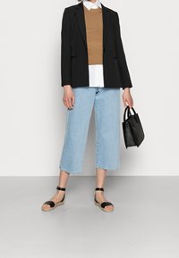 Blazer noir sur un pull en tricot beige, chemise blanche en dessous, jean droit bleu clair, et sandales à bride noire; petit sac à main noir.