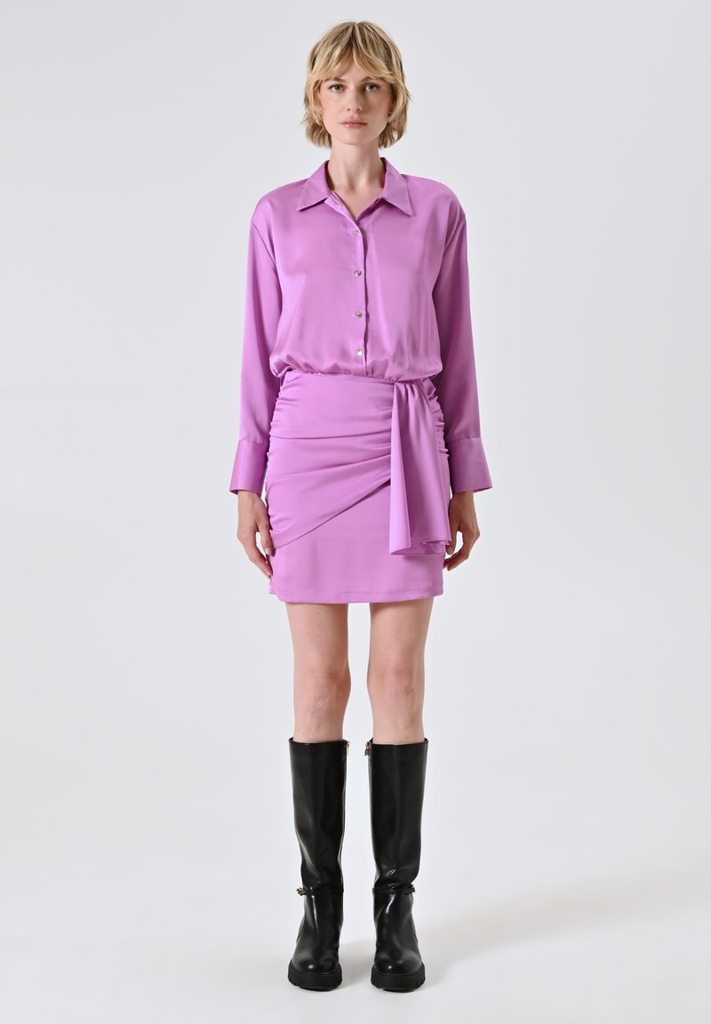 Lavendelfarbenes Seidenhemd mit Knöpfen, kombiniert mit einem drapierten lavendelfarbenen Minirock mit einer gebundenen Akzentuierung. Schwarze knielange Stiefel vervollständigen den Look.