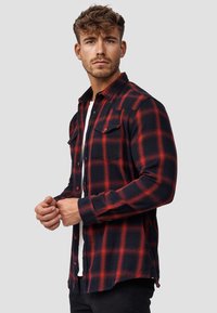 Camisa de franela a cuadros rojos y negros, de manga larga, con botón en el frente, dos bolsillos en el pecho, tela texturizada. Usada sobre una camisa blanca.