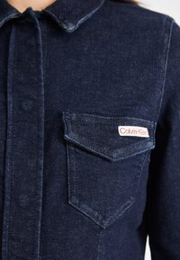 Mørkeblå denimjakke med krage, foran lommer og et rødt Calvin Klein-logo på venstre lomme. Glatt tekstur, standard passform.
