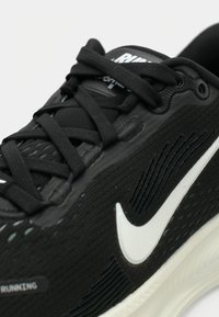 Zapatilla de correr negra con parte superior texturizada, swoosh blanco de Nike y cordones negros planos. Diseñada para ofrecer comodidad y soporte con una suela acolchada.