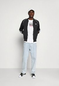 Black bomber jacket with white stripes, white t-shirt with graphic, light blue jeans, and black sneakers with white accents, person standing.