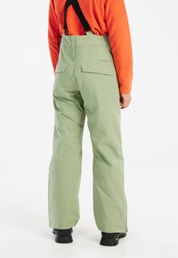 Groene waterdichte sneeuwbroek met verstelbare zwarte banden, een soepele textuur, twee achterzakken en een relaxte pasvorm, gecombineerd met een oranje top.