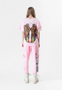 Camiseta rosa con alas bordadas coloridas en la espalda, combinada con pantalones rosa a juego que presentan patrones de plumas blancas y etiquetas.
