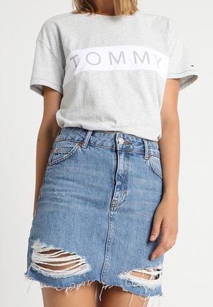 Vrouw draagt een grijs T-shirt met 'TOMMY' opgespeld, ingestopt in een hooggeplaatste blauwe denim rok met twee gescheurde details vlak bij de zoom.