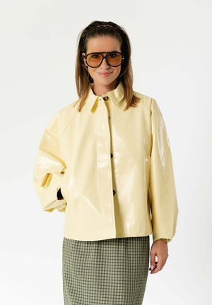 Femme portant une veste jaune pâle surdimensionnée, de grandes lunettes teintées et une jupe à carreaux verte, debout avec une main dans la poche.