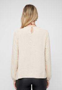 Beige langslanke top met textuurafwerking, featuring een keyhole rugdetail en elastische manchetten. De zoom is aan de achterkant iets langer.