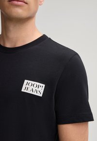 Zwarte t-shirt met korte mouwen, ronde halslijn en een wit rechthoekig logo-embleem met de tekst "JOOP! JEANS" op de linkerkant van de borst.