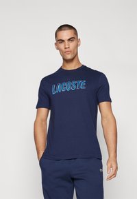 Mornarsko modra majica s kratkimi rokavi, s klasičnim ovratnikom, izdelana iz bombaža, na sprednji strani pa je v svetlo modri barvi jasno napisan napis "LACOSTE".