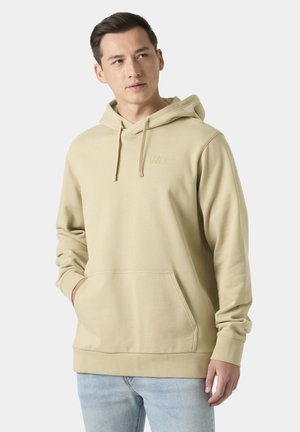 Joven con sudadera con capucha beige con bolsillo frontal y vaqueros azul claro, mirando ligeramente hacia un lado contra un fondo liso.