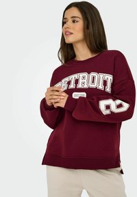 Burgundin sydänpaita, jossa on teksti "DETROIT" ja numero "2" hihoissa. Ominaisuuksiin kuuluvat pyöreä kaula-aukko, ribbaudet hihansuissa ja sivuaukot helmassa.