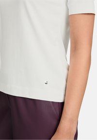 Haut blanc à manches courtes avec un ourlet arrondi, présentant un accent métallique près du bas, associé à un pantalon en cuir violet.
