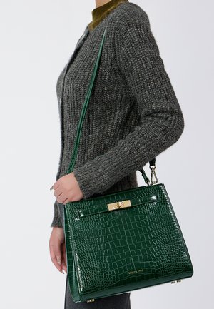 Persoon in een grijze gebreide trui die een donkergroene krokodillenleder handtas vasthoudt met een gouden sluiting en schouderband.