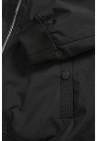 Manche de veste noire avec poignet côtelé, fermeture éclair métallique et détail de poche à bouton-pression sur tissu lisse.