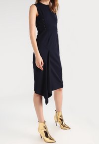 Mörkblå ärmlös klänning med asymmetrisk fåll, knappar i sidan och en slank siluett, kombinerad med glänsande guldfärgade ankelboots.