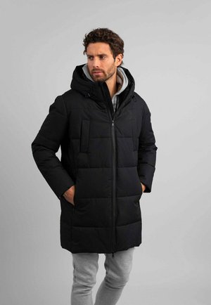 Homme portant un manteau matelassé noir à capuche par-dessus un sweat à capuche gris et un pantalon gris, mains dans les poches du manteau, regardant sur le côté.