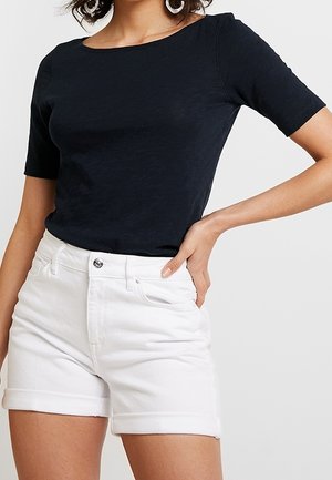 Vrouw die een zwart kortermig topje en een witte denim short met opgerolde boorden draagt, poserend tegen een effen achtergrond.