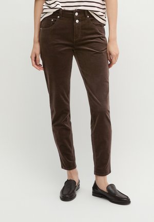 Person trägt dunkelbraune Slim-Fit-Hose, schwarze Slipper und ein eingestecktes gestreiftes Oberteil, steht vor einem einfarbigen hellen Hintergrund.
