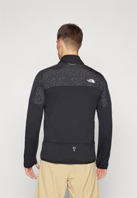 Melna zip-up jaka ar gludu tekstūru, ar rakstainu augšējo daļu un logotipu kreisajā plecā. Regulējams apkakles dizains.