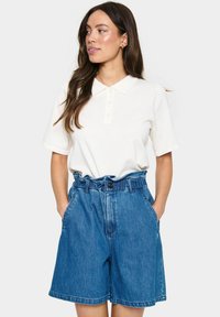 Weißes Poloshirt mit kurzen Ärmeln, Kragen und Knopfleiste, kombiniert mit hochgeschnittenen blauen Jeansshorts mit einem Taillengürtel und seitlichen Taschen.