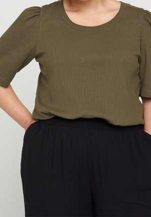Personne portant un haut côtelé vert olive à manches courtes et un pantalon noir ample, les mains dans les poches sur un fond uni.