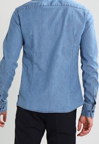 Ljusblå denimskjorta med lång ärm, skräddarsydd passform, subtila sömnadsdetaljer och knäppte ärmslut. Slät textur och rundad fåll.