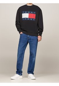 Svart sweatshirt med vitt, rött och blått "TOMMY JEANS"-logotyp, ihop med blå denimjeans och vita sneakers. Avslappnad passform och texturerade tyger.