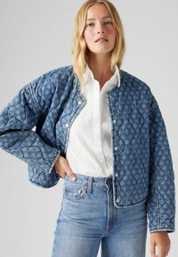 Kvadrovana modra denim jakna z pritisnimi gumbi, teksturiranimi valovitimi vzorci in sproščenim krojem. V kompletu z belo srajco s gumbi in modrimi kavbojkami.