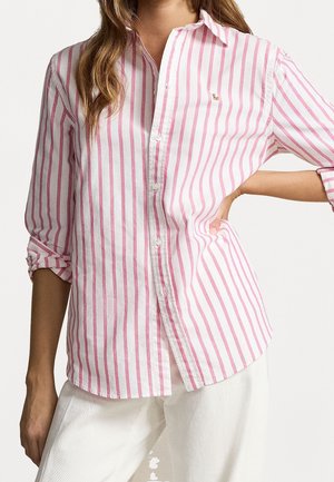 Kobieta ubrana w różowo-białą koszulę zapinaną na guziki z pionowymi paskami, z podwiniętymi rękawami, w połączeniu z białymi spodniami.
