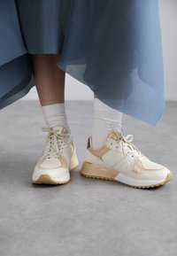 Witte en beige sneakers van mesh en leer, veterontwerp, gouden accenten en getextureerde zolen, gecombineerd met witte sokken en een blauwe rok.