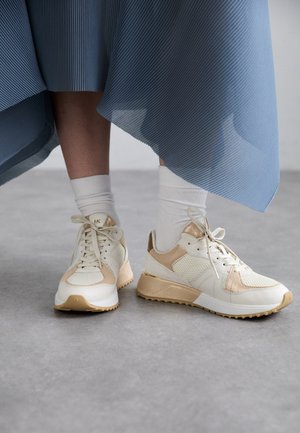 Baskets blanches et beiges en matériaux mesh et cuir, à lacets, avec des accents dorés et des semelles texturées, portées avec des chaussettes blanches et une jupe bleue.