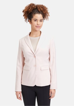 Hellrosa maßgeschneiderte Blazerjacke mit Revers, zwei vorderen Taschen und Knöpfen. Kombiniert mit einem beigen Strickoberteil und dunklen Hosen.
