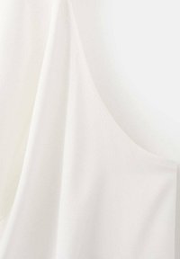 Top de tela blanca con una textura suave, que presenta líneas curvas y una silueta amplia para un ajuste relajado, enfatizando el diseño minimalista.