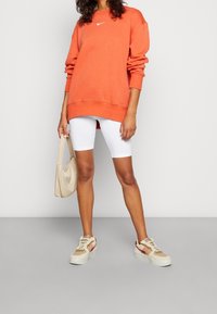 Nike orange sweatshirt med en vit logotype, vita cykelshorts och beige sneakers med rosa detaljer. Håller en ljusbeige väska.