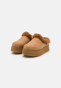 Tan suede klogger med en fluffy brun pelskant og en kilehæl, med en teksturert overflate og sømdetaljer langs kanten.