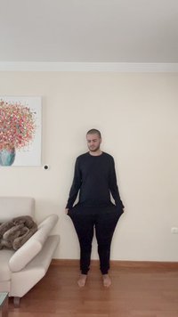 Camisa negra de manga larga y pantalones negros holgados, de pie descalzo, con tela cayendo alrededor de las piernas y los brazos en un entorno interior.