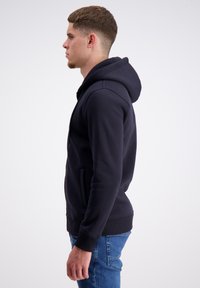 Mørkeblå hoodie lavet af bomuldsblanding med en frontlomme, ribbet manchet og kant, og med hætte der har snører. Modellen vises i profil.