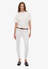 Una camiseta blanca de algodón combinada con unos jeans skinny blancos, un cinturón de cuero trenzado marrón y mocasines oscuros, posando contra un fondo sencillo.
