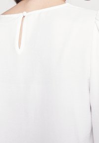 Vue arrière d'un chemisier blanc avec un col rond, une ouverture en forme de trou de serrure et une fermeture par un seul bouton à la nuque.