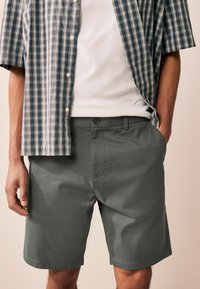 Grau Shorts mit Seitentaschen, glatter Textur und Knopfverschluss. Kombiniert mit einem kurzärmligen blau-weißen karierten Hemd.