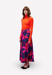Langärmliges orangefarbenes Oberteil, kombiniert mit einem fließenden, gemusterten Rock mit pinken und blauen Blumenmustern. Schwarze Schuhe runden das Outfit ab.