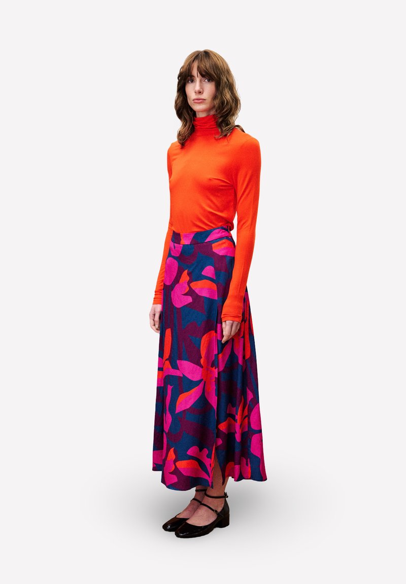 Langärmliges orangefarbenes Oberteil, kombiniert mit einem fließenden, gemusterten Rock mit pinken und blauen Blumenmustern. Schwarze Schuhe runden das Outfit ab.