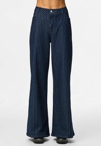 Tumbra denim bikses tumši zilonī ar baltām vertikālām svītrām, kas aprīkotas ar priekšējo pogu aizdari un diviem sānu kabatām. Gluda tekstūra.