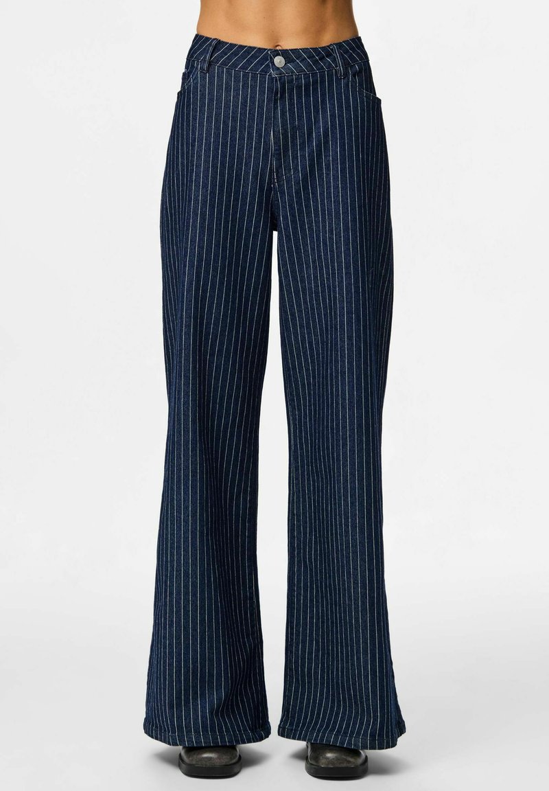 Tumbra denim bikses tumši zilonī ar baltām vertikālām svītrām, kas aprīkotas ar priekšējo pogu aizdari un diviem sānu kabatām. Gluda tekstūra.