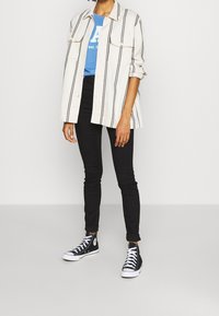 Layered outfit featuring a white and black striped button-up shirt, blue graphic t-shirt, black skinny jeans, and black high-top sneakers.