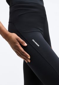 Κοντινή λήψη ενός χεριού που ακουμπάει πάνω σε μαύρο αθλητικό κολάν με ορατό το λογότυπο της Salomon στον μηρό, σε λευκό φόντο.