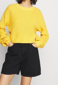 Suéter amarillo de punto con textura acanalada y hombros caídos, combinado con pantalones cortos de mezclilla negros de tiro alto con bolsillos laterales.